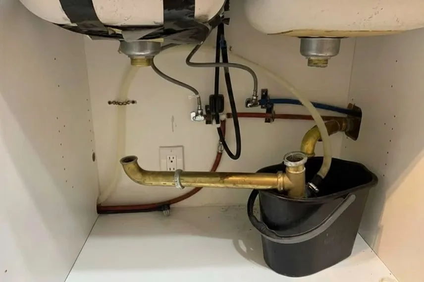 Old metal pipes under a sink with a bucket collecting water
